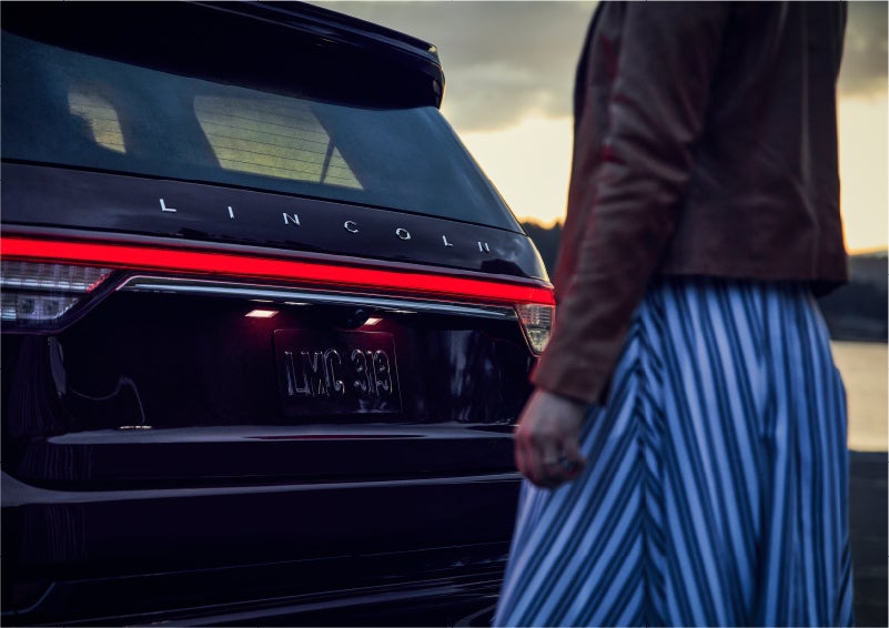 A person is shown near the rear of a 2023 Lincoln Aviator® SUV as the Lincoln Embrace illuminates the rear lights | Bondy's Lincoln in Dothan AL